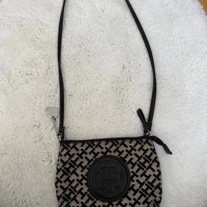 vintage Tommy Hilfiger svart axelväska - Snygg äkta vintage axelväska från Tommy Hilfiger med svart och beige mönster, rund TH-logga framtill och silverfärgad tag. Väskan har justerbar axelrem, dragkedja och innerfack. Perfekt storlek för det viktigaste och en stilren detalj till din outfit.