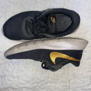 Svarta Nike sneakers med guld swoosh - Nike sneakers i svart mesh med en snygg guld swoosh på sidan och svart snörning. Skorna har en vit, mönstrad sula och klassisk Nike-logga på plösen. Perfekta för dig som gillar sportig och stilren vibe. Väl använda. 