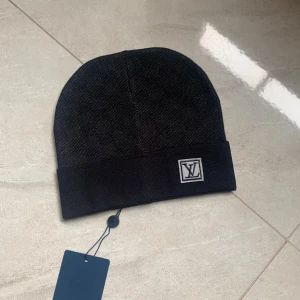 Svart mössa från Louis Vuitton - Stilren svart mössa från Louis Vuitton med diskret LV-logga framtill. Mössan är ribbstickad med uppvikt kant och har en exklusiv känsla. Perfekt accessoar för att lyfta din look under kyliga dagar.