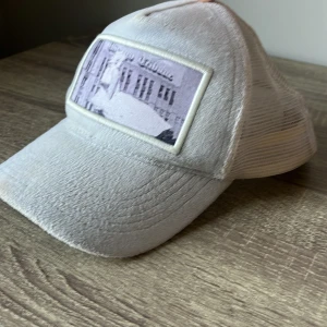 Emilio pendenza keps - Snygg vit keps med mesh på baksidan och ett coolt tryck framtill. Framsidan är i mjukt tyg och har en rektangulär patch med motiv i grå toner. Perfekt för dig som gillar streetstyle och vill ha en luftig keps till sommaren.