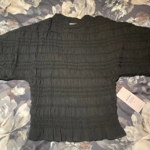 Svart plisserad blus från Nelly - Säljer en svart blus från Nelly i storlek XS. Blusen har plisserad struktur med breda, korta ärmar och rundad halsringning. Materialet är lätt och ger en luftig känsla, perfekt för att skapa en cool och trendig look.