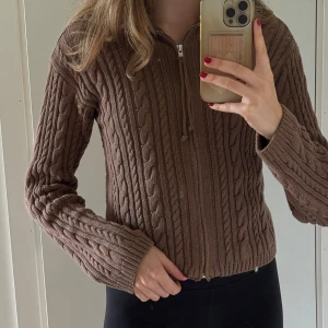 Brun stickad kofta från Djerf Avenue - Säljer en brun stickad kofta från Djerf Avenue med kabelstickat mönster och dragkedja framtill. Koftan har hög krage och långa ärmar. Supermysig och snygg till både jeans och kjol. Köpt för 1500.