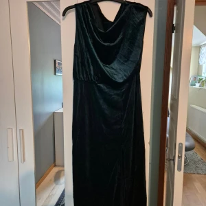 Mörkgrön sammetsklänning med drapering - Elegant långklänning i mörkgrön sammet med snygg draperad halsringning. Klänningen är ärmlös och har en mjuk, lyxig känsla som verkligen sticker ut. Perfekt för dig som vill ha en klassisk och stilren look med lite extra edge.