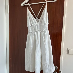 Vit kort sommarklänning från Shein - Supersöt vit klänning från Shein i storlek M. Den har smala axelband, v-ringad front och smockad midja med volangdetaljer. Klänningen är kort och gjord i ett lätt bomullstyg, perfekt för varma dagar. Snyggt kors i ryggen och luftig passform. Liten fläck på framsidan men inget jag personligen tycker att man märker av (se bild)