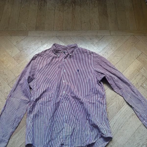 Rödvit randig skjorta Polo Ralph Lauren - Snygg skjorta från Polo Ralph Lauren i rödvitt randigt mönster. Klassisk krage, knäppning framtill och lång ärm. Broderad logga i blått på bröstet. Perfekt för dig som gillar stilrena och tidlösa plagg.