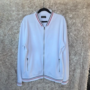 Vit tröja med dragkedja från Zara Man - Vit tröja från Zara Man med dragkedja framtill och ribbade muddar med röda och svarta ränder. Tröjan har två fickor med dragkedja.