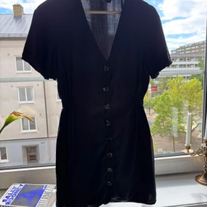 Svart kort klänning från Zara - Svart kort klänning från Zara med v-ringning, knappar längs hela framsidan och korta ärmar. Klänningen har en lätt insvängd midja och är tillverkad i ett tunt, mjukt tyg som känns luftigt. Perfekt för dig som gillar enkel och stilren design.