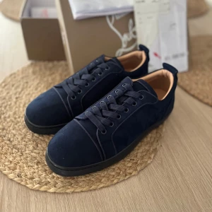 Mörkblå sneakers från Christian Louboutin 42! - Snygga mörkblå sneakers från Christian Louboutin i mocka med klassisk röd sula och diskret broderad logga på hälen. Skorna har rund tå, svarta snören och en stilren design som sticker ut med den ikoniska röda undersidan.