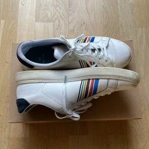 Vita sneakers från Paul Smith med ränder - Snygga vita sneakers från Paul Smith med färgglada ränder i blått, rött, grönt och gult på sidorna. Skorna har snörning, rund tå och platt sula. Ovandelen är i skinn och hälen har en svart detalj. Perfekta för en stilren och färgglad look. Helt nya snören.