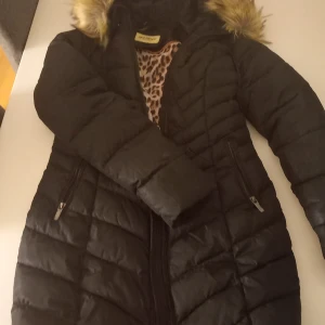 Svart lång pufferjacka Roco Baroco - Svart lång pufferjacka från Roco Baroco med snygg quiltad design och fuskpäls på huvan.  Finns även ett skärp till. Jackan har dragkedja framtill och två praktiska fickor med dragkedja. Insidan har ett coolt leopardmönster som ger extra edge till looken.