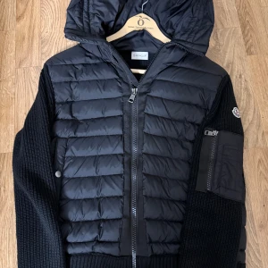 Moncler cardigan  - Påse och andra grejer medföljer.Svart pufferjacka från Moncler med stickade ärmar och quiltad front. Jackan har huva, dragkedja framtill, knappdetaljer och en ficka med dragkedja på ärmen. Materialmix av dun, polyester och stickat ger en schysst look. Perfekt för dig som gillar stilren design och premiumkänsla. Den är ny har haft den ungefär 2 månader inga problem med den. Påse och andra grejer medföljer!!!! Storlek S material akp!!!!