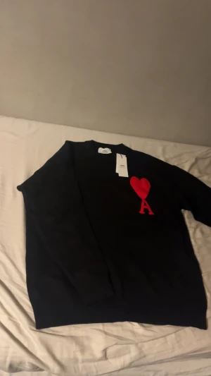 Svart stickad tröja från Ami med hjärta - Svart stickad tröja från Ami med rund hals och lång ärm. På bröstet finns ett stort rött hjärta med bokstaven A, vilket ger en unik och trendig vibe. Ribbstickade muddar vid ärmslut och nederkant. Perfekt för dig som gillar stilrena detaljer.
