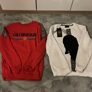 Glorious Gangsta sweatshirts i rött & vitt - Två sweatshirts-tröjor från Glorious Gangsta, en röd med svarta och guldiga detaljer samt tryck på bröstet och mönstrade band på ärmarna, och en vit med svart pantermotiv och liknande banddetalj. Båda har lång ärm och är i mjuk bomull. 200kr st eller 300kr för båda