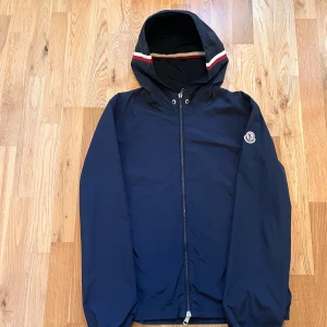 Mörkblå vindjacka från Moncler - Snygg mörkblå vindjacka från Moncler med huva och coola rödvita detaljer på kanten. Jackan har dragkedja framtill, lång ärm och Moncler-logga på armen. Materialet är lätt och känns sportigt, perfekt för blåsiga dagar. Stängning med dragkedja och tryckknappar vid halsen. Storlek 1 vilket motsvarar storlek S!