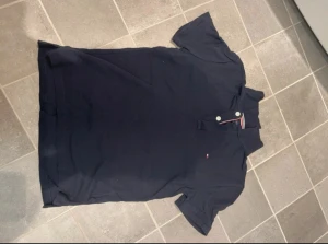 Mörkblå pikétröja Tommy Hilfiger - Snygg mörkblå pikétröja från Tommy Hilfiger med klassisk krage och två vita knappar framtill. Diskret logga broderad på bröstet och coola ränder i rött, vitt och blått på insidan av kragen. Perfekt för en clean och sportig look.