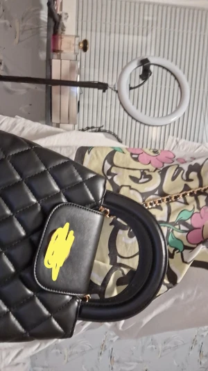 Svart quiltad handväska med kedja - Snygg svart handväska i quiltad design med guldfärgade detaljer och kedjerem. Väskan har rundade handtag och ett lock med vridlås. Tillverkad i skinnimitation och passar perfekt för dig som gillar en klassisk men trendig look.