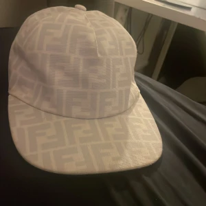 Vit Fendi keps med logomönster - Snygg vit keps från Fendi med det klassiska FF-logomönstret över hela kepsen. Kepsen har en rak skärm och justerbar baksida. Tillverkad i 100% polyester, perfekt för dig som gillar exklusiva accessoarer med stilren design.