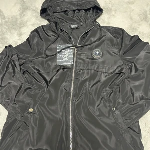 Svart vindjacka från Trapstar S - Svart vindjacka från Trapstar med huva och dragkedja framtill. Jackan har snygga detaljer som logopatch på bröstet, snörning vid huvan och praktiska fickor med knappstängning. Materialet är glansigt och lätt, perfekt för streetwear.