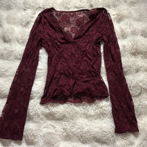 Vinröd spetsblus med v-ringning - Säljer en vinröd blus i transparent spets med blommigt mönster. Blusen har långärmade, lätt utsvängda ärmar och en djup v-ringning. Materialet är mjukt och stretchigt, perfekt för dig som gillar romantisk och edgy stil. Men har ett litet hål i sig längst ner på tröjan 