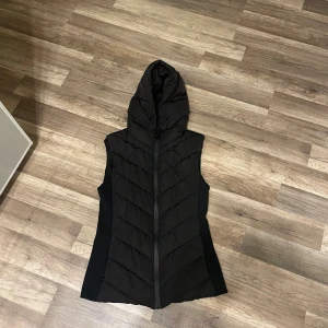 Svart dunväst med huva från Zara - Snygg svart dunväst från Zara i storlek S. Västen har en quiltad design med dragkedja framtill och en stor huva med snörning. Perfekt för lager-på-lager och har en stilren, lite längre passform som ger en cool look.