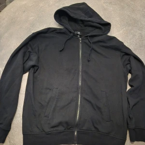 Svart hoodie med dragkedja från H&M - Svart hoodie från H&M med huva och snörning, hel dragkedja framtill och två fickor. Tröjan är långärmad och har ribbade muddar vid ärmslut och nederkant.