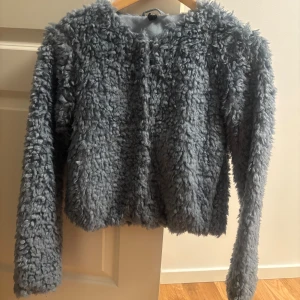 Blå teddyjacka från H&M Divided XS - Säljer en fluffig blå teddyjacka från H&M Divided i storlek XS. Jackan har långärmade ärmar, rund hals och stängs med diskreta knappar framtill. Perfekt för dig som gillar mysiga och trendiga jackor med cool textur.