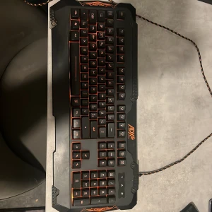 ADX Ultimate Gaming Keyboard - Snyggt ADX Ultimate Gaming tangentbord med orange bakgrundsbelysning och robust design. Fullstort med numerisk del, flätad kabel och nordisk layout. Tangenter och yta ser ut att vara i gott skick utan synligt slitage. Perfekt för gaming och produktivitet.