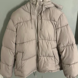 Hm jacka  - Säljer en beige pufferjacka från H&M i storlek XS. Jackan har en stor, mysig huva, dragkedja framtill och snörning vid halsen. Den är quiltad och har två fickor på sidorna. Perfekt för kalla dagar och har en loose passform.