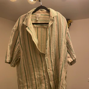 Skjorta från H&M - Oversized skjorta från H&M i beige med gröna ränder. Skjortan har korta ärmar, klassisk krage och knappar framtill. 