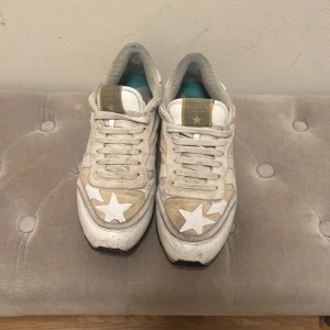 Valentino Garavani sneakers med stjärnor - Beiga och vita sneakers från Valentino Garavani med coola stjärndetaljer i vitt på sidorna och tån. Baksidan har en stjärna med nitar och blå sten, samt rockstuds runt hälen. Skorna har snörning, platt sula och är tillverkade i mocka och skinn. Perfekt för dig som gillar unik design.