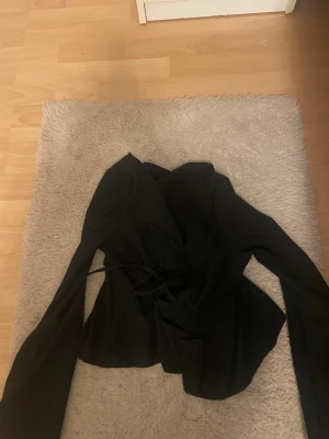 Svart omlott bolerotopp med knytband - Säljer en svart bolerotopp med långa ärmar och snygg omlott-design. Toppen har knytband i midjan för en justerbar passform och är croppad i modellen. Perfekt för att lyfta vilken outfit som helst med en stilren touch.