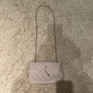 Vit quiltad axelväska med kedja - Supersöt vit axelväska i quiltad design med guldfärgad kedja och ett kronformat spänne med pärldetalj. Väskan är gjord i syntetmaterial och har svart innerfoder. Perfekt för dig som gillar stilrena och eleganta accessoarer.