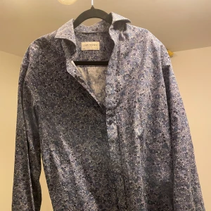 Skjorta från John Henric - Snygg skjorta från John Henric med blommigt mönster i olika blå nyanser och inslag av vitt. Skjortan har klassisk krage, knappar framtill och lång ärm. Sitter som en större M eller mindre L