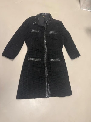 Svart klänning från Maje, strl 40 - Snygg svart klänning från Maje i storlek 40. Klänningen har en cool quiltad struktur och glansiga detaljer längs midjan och fickorna. Materialet är en mix av polyester och bomull, vilket gör den både bekväm och stilren. Perfekt för dig som gillar en edgy och modern look.