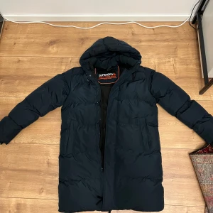 Mörkblå lång pufferjacka Superdry - Säljer en mörkblå helt Ny Oanvänt lång pufferjacka från Superdry med huva och broderad logga på ärmen. Jackan har dragkedja framtill, två sidofickor och är fodrad med svart innerfoder. Perfekt för kalla dagar och har en clean, sportig look.