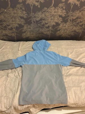 Blå och grå vindjacka från Under Armour - Snygg tvåfärgad vindjacka från Under Armour i ljusblått och grått. Jackan har huva, hel dragkedja framtill och två sidofickor. Perfekt för dig som gillar sportig stil och vill ha något lätt och funktionellt. Logga på bröstet och stilren design.