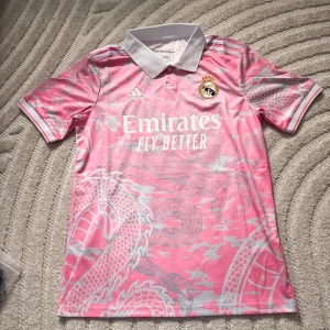 Rosa Real Madrid fotbollströja Adidas - Snygg rosa Real Madrid fotbollströja från Adidas med grått drakmönster och vit krage. Tröjan har korta ärmar, klubbmärke på bröstet och tryckta sponsorloggor. Perfekt för dig som vill sticka ut på planen eller på läktaren.