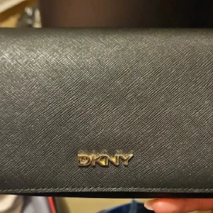 Svart plånbok från DKNY i skinn - Snygg svart plånbok från DKNY med stilren design och guldfärgad logga framtill. Plånboken är tillverkad i strukturerat skinn och har en klassisk, avlång form som känns både lyxig och tidlös. Perfekt för dig som gillar minimalistiska accessoarer.