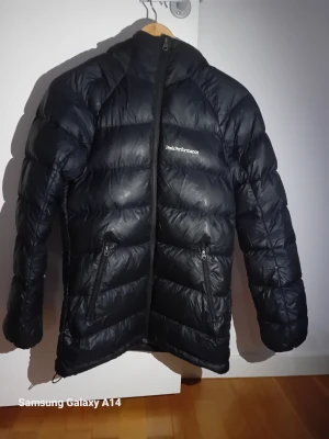 Svart Peak Performance dunjacka XS - Säljer en svart dunjacka från Peak Performance i storlek XS. Jackan har huva, två sidofickor med dragkedja och ett stilrent, puffigt quiltat mönster. Perfekt för kalla dagar och har en snygg, sportig look med logga på bröstet.
