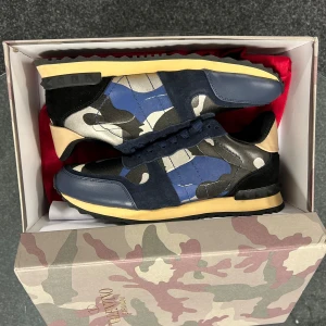 Valentino Garavani Rockrunner sneakers - Snygga Valentino Rockrunner sneakers med blått och beige camouflage-mönster, svarta och mörkblå detaljer i mocka och skinn. Platt sula med nitar och rund tå. Ikonisk design som sticker ut med coola färgblock och robust yttersula. Det står storlek 41 men dom sitter så att de passar någon med storlek 43-44