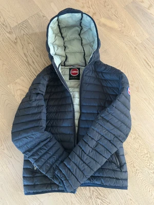 Mörkblå Colmar pufferjacka med huva - Snygg mörkblå pufferjacka från Colmar med huva och coola logopatchar på ärm och nederkant. Jackan har quiltad design, dragkedja framtill och två sidofickor med zip. Insidan är ljusgrå vilket ger en schysst kontrast. Perfekt för dig som gillar street och sportig stil.