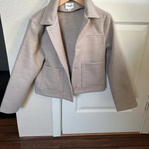 Beige overshirt/jacka från Kappahl - Snygg beige overshirt/javka från Kappahl med klassisk krage, stora framfickor och knappar framtill. Tyget är polyester och viskos, perfekt för lager-på-lager och chill stil. Passar dig som gillar enkel och clean look.