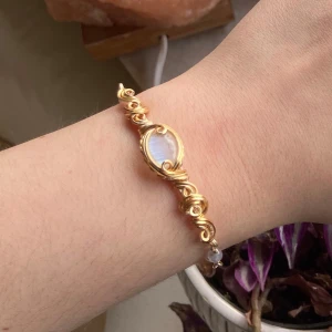 Guldfärgat armband med månsten - Unikt armband med guldfärgad kedja och handgjorda snirkliga detaljer runt en oval skimrande månsten i mitten. Armbandet har små pärlliknande vita stenar och justerbar kedja för perfekt passform. Perfekt för dig som gillar boho och magiska vibes.