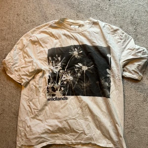 Vit t-shirt med blommigt tryck från H&M - Säljer en vit t-shirt från H&M i relaxed fit med stort svartvitt blommigt tryck och texten 'wildlands' på framsidan. Baksidan har tryckt text i svart.