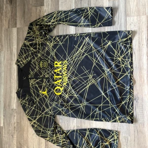 PSG långärmad träningströja svart/gul - Snygg långärmad fotbollströja från PSG i svart med gula grafiska linjer och klubbmärke på bröstet. Tröjan har dragkedja vid halsen och tryck med 'Qatar Airways' samt 'Paris' på ärmen. Tillverkad i glansigt, lätt funktionsmaterial.