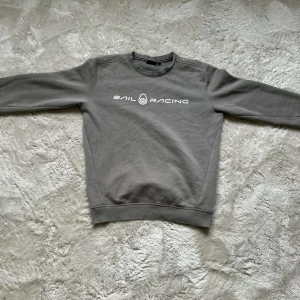 Grå Sail Racing sweatshirt i bomull - Snygg grå sweatshirt från Sail Racing med vit logotyp tryckt på bröstet. Tröjan har rund hals, ribbade muddar och är tillverkad i mjuk bomull. Perfekt för dig som gillar stilren och sportig look.
