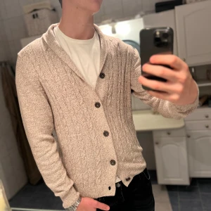 Beige cardigan - Snygg beige kofta i storlek M. Koftan har kabelstickat mönster, ribbade muddar och fyra mörka knappar framtill. Den har en skön krage och är långärmad, perfekt att slänga på över en t-shirt. Materialet är stickat och känns mjukt mot huden. 