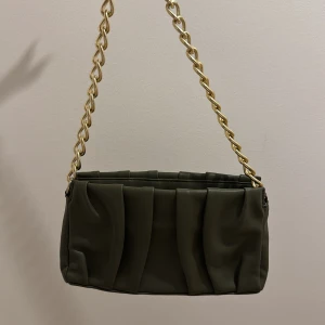 Grön axelväska med kedja - Stilren grön axelväska i syntetmaterial med veckad design och bred guldfärgad kedja som axelrem. Väskan har dragkedja upptill och en modern, rektangulär form som ger en cool vibe. Perfekt för dig som vill sticka ut med accessoarer.
