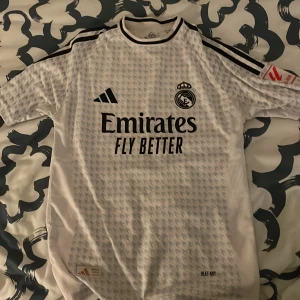 Real Madrid Mbappé #9 fotbollströja - Säljer en vit Real Madrid fotbollströja från Adidas med Mbappé och nummer 9 på ryggen. Tröjan har korta ärmar, svarta detaljer och ett subtilt randigt mönster. Tryck från La Liga och HP på ärmarna samt Emirates Fly Better på bröstet. Materialet är lätt och andas. Priset kan diskuteras 