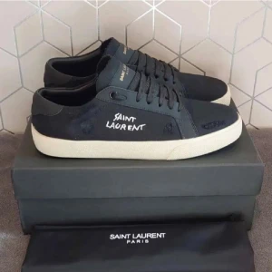 Svarta Saint Laurent sneakers - Svarta sneakers från Saint Laurent med vit handskriven logga på sidan. Skorna har en klassisk låg profil, snörning och beige platt gummisula. Ovandelen är i canvas och detaljer i skinn. Perfekta för en clean och stilren look.